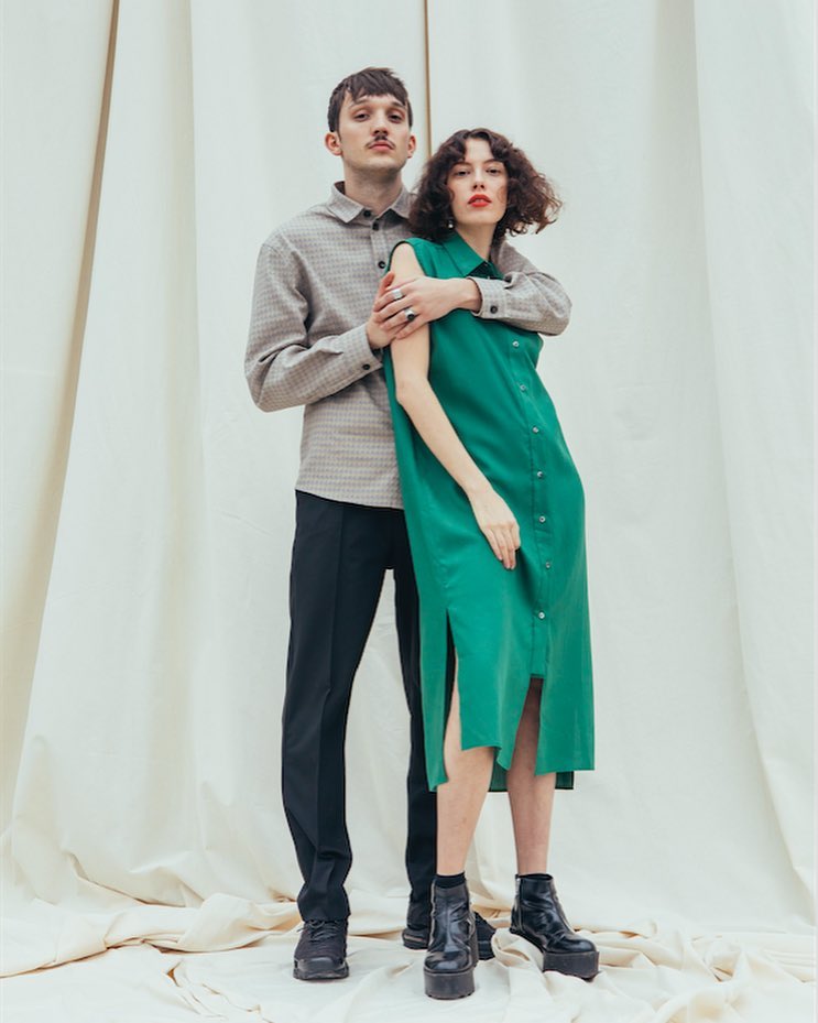 Male and female models wearing a grey button-up shirt, black pants and a green button-up sleeveless dress from HUNDHUND with black shoes