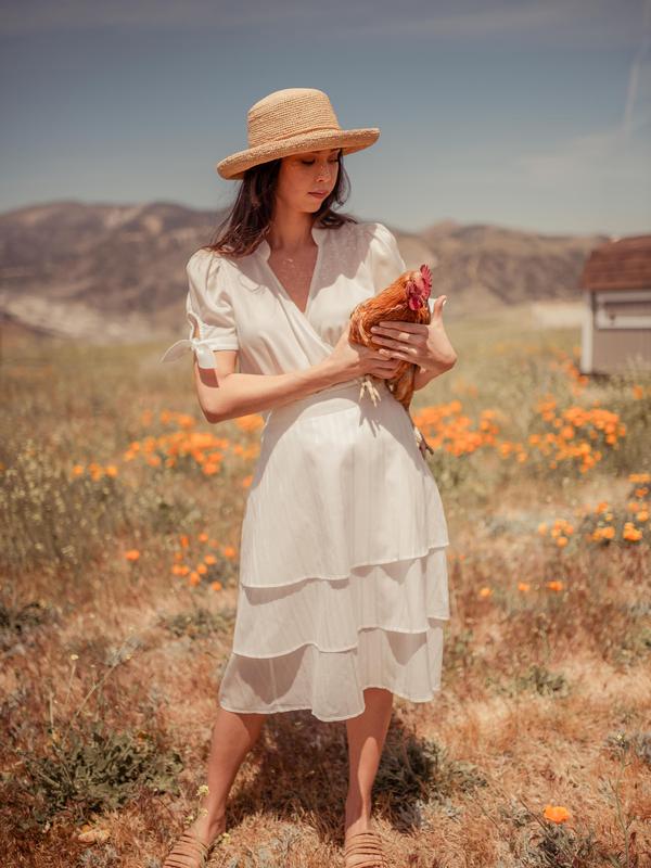 Model wearing white shirt and skirt from Valani with a straw hat in a field while holding a chicken
