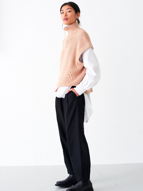Model wearing black trousers with a beige turtleneck sweater vest and a white button-up shirt underneath from sustainable brand Mother of Pearl