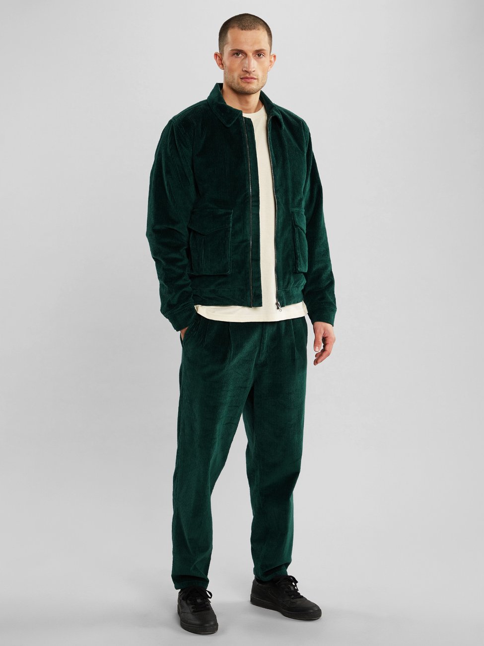 Male model wearing dark green corduroy pants with a matching jacket and a white top underneath from Dedicated