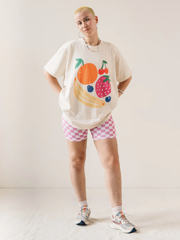 Model wearing an off-white oversized T-shirt with drawings of fruits on it, and pink and white checkered bike shorts from sustainable brand Nube