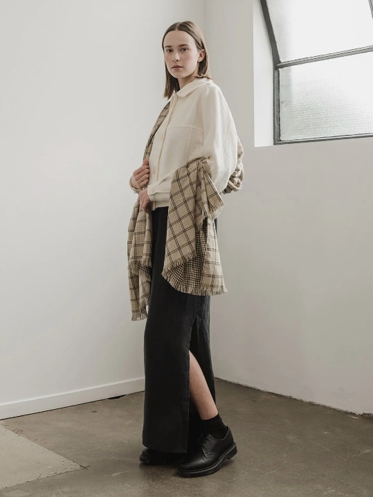 Female model wearing a long black shirt and a white jacket and a beige plaid scarf from sustainable brand A.BCH with black shoes