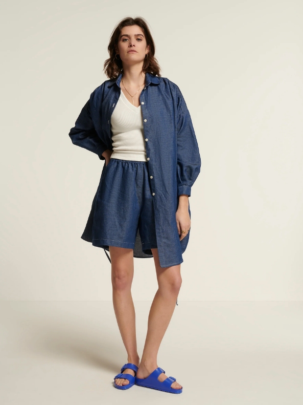 Model wearing dark blue shorts, a matching long button-up shirt and a white top from sustainable brand New Optimist