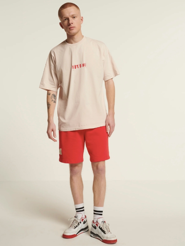 Male model wearing red shorts and a beige T-shirt from sustainable brand New Optimist