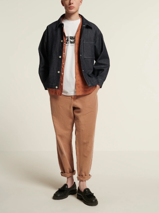 Male model wearing beige pants, a dark grey denim jacket, a coral coloured button-up shirt and a white T-shirt from sustainable brand New Optimist