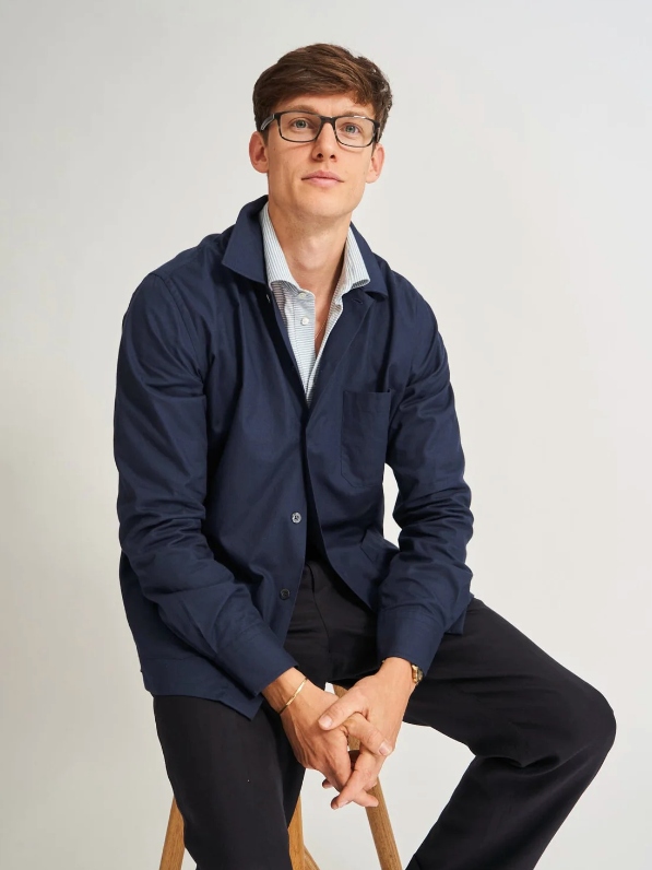 Male model wearing black pants, a light blue button-up shirt and a dark blue overshirt from sustainable brand NEEM