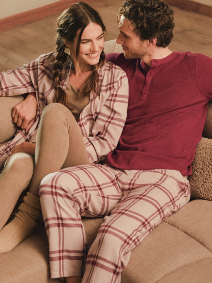 A male and female model both wearing checked red and beige loungewear/sleepwear from sustainable brand PACT, while sitting on a couch